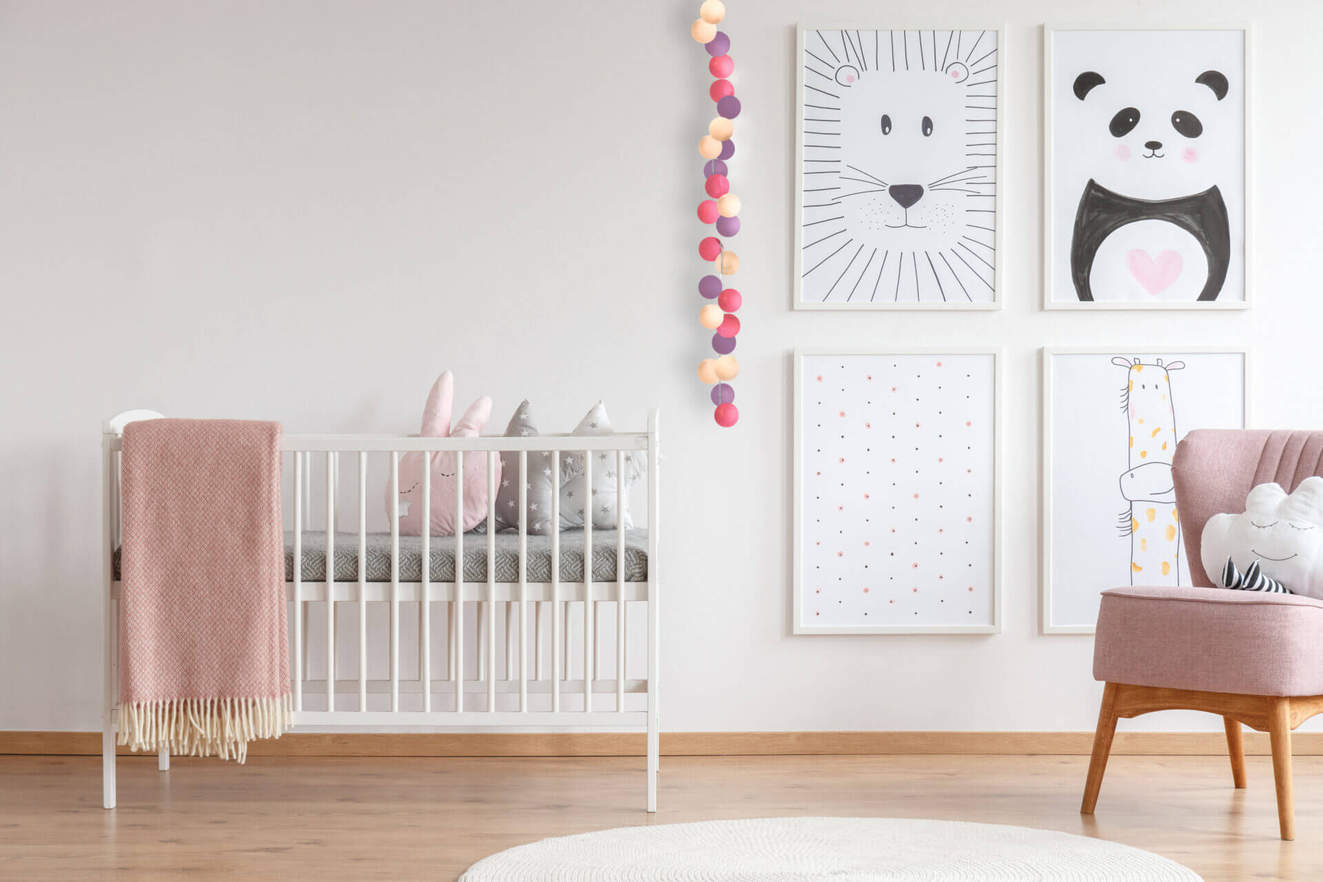 White crib with pink blanket and decorative cushions standing in cute baby room with posters