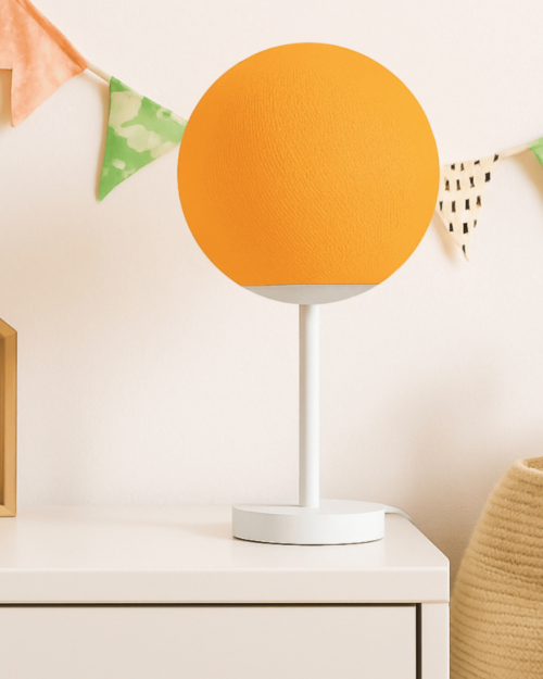 Orangefarbene Tischlampe mit handgefertigter Baumwollkugel auf weißem Standfuß; warmweißes LED-Nachtlicht auf dem Nachttisch im Kinder- und Jugendzimmer.